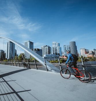 A cyclist rides across a modern bridge with a city skyline in the background, under a clear blue sky.